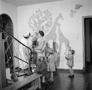 Erzieherin in einer Kinderkrippe (oder einem Kindergarten) mit Kindern und einer Mutter, die ihren kleinen Sohn auf dem Arm trägt