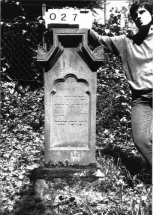 Grabstein 27 (Index: 100101186) Material: Sandstein Zustand: am Sockel verwittert Maße HxBxT: 133x49x26 cm Beschreibung: Stele, profilierter Sockel, eingezogener giebelartiger Abschluß rechteckig erhöht mit Verdachgung und Eckakroteren, Schriftfeld mit Rundbogenabschluß Symbolik: Rundbogenfries um das Schriftfeld, Blätter in den Akroteren: Grabstein 27