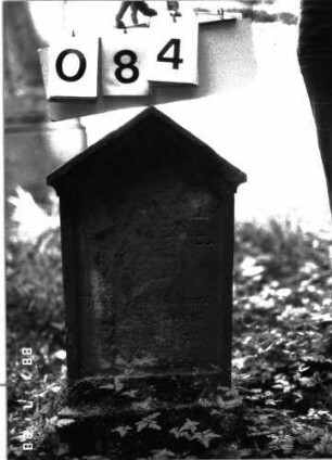 Grabstein 84 (Index: 100101300) Material: Sandstein Zustand: stark abgeblättert mit großem Textverlust Maße HxBxT: 70x33x12 cm Beschreibung: Stele, Sockel, giebelartiger Abschluß mit Verdachung Besonderheit: Kindergrab: Grabstein 84