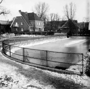 Ziegeleistraße: vorn Löschteich mit Einfriedung und Schild: dahinter Bauernhäuser: im Hintergrund Leuchtreklame der Ziegelwerke H. Schlick: Winter