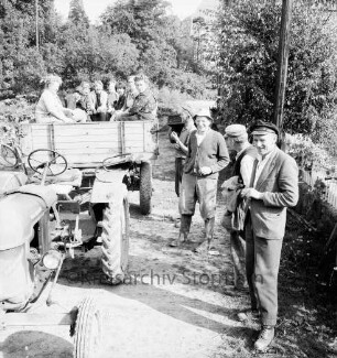 Trecker und Anhänger mit Kartoffelerntehelferinnen: vorn rechts Landwirte und Erntehelfer: dahinter Bäume