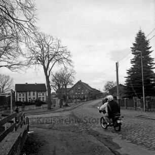 Dorfstraße: Kreisstraße zwischen Bad Oldesloe und Tremsbüttel: Kopfsteinpflaster und Schlaglöcher: links Abzweigung der Lindenstraße mit Bäumen: vorn rechts Motorrad mit zwei Personen: im Hintergrund Bauernhäuser und Wohnhaus