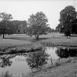 Schlosspark: äußerer Schlossgraben mit Seerosen, Schwan: Angler: Spaziergänger auf Fußweg, Parkbank, Bäume: im Hintergrund Gottesbuden
