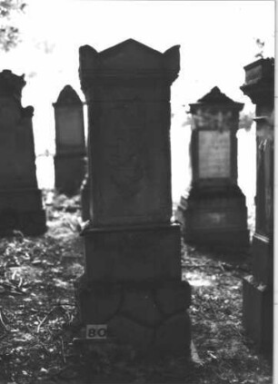Grabstein 80 (Index: 99898841) Material: Sandstein Zustand: stark verwittert Maße HxBxT: 169x54x26 cm Beschreibung: aufgesockelte Stele, Giebelaufsatz mit Eckakroterien, unterer Sockel felsartig gearbeitet: Grabstein 80