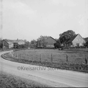 Kreisstraße aus Richtung Stubben (Kreis Segeberg): dahinter Zaun und Weide: im Hintergrund Ortseingang mit Bauernhäusern