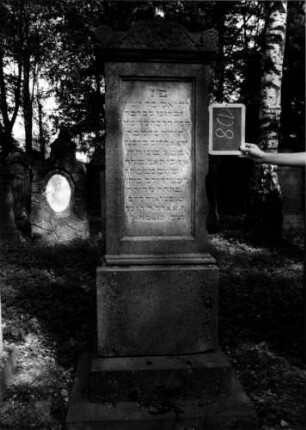 Grabstein 128 (Index: 100111262) Material: Sandstein Zustand: leicht verwittert mit Textverlust, beschädigt Maße HxBxT: 194x60x49 cm Beschreibung: Stele, getreppter Sockel, Abdeckplatte mit Volutenaufsatz, rechteckige Schriftfelder eingetieft Symbolik: Blüten, Voluten: Grabstein 128