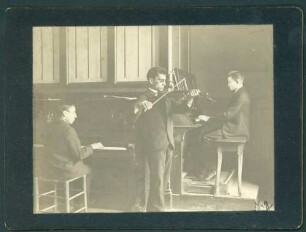 Trio (Klavier, Geige, Harmonium) musizierender Männer.
