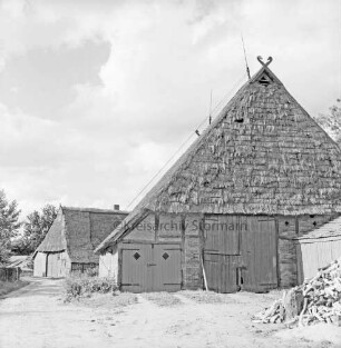 Bauerngehöft mit zwei Reetdachscheunen. vorn rechts Stapel mit Holzscheiten: hinten links Bäume