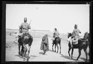 Nador (Marokko): 3 berittene Offiziere [des spanischen 8. Kavallerie-Regiments "Lusitania"] in hellgestreiften Uniformhosen, dunklen Uniformjacken und Tschakos (=militärische Kopfbedeckung in zylindrischer oder konischer Form) bei einem Einheimischen, im Hintergrund Gebäude (Rückansicht) ("Gefangener Mörder auf dem Transport von Nador nach Melilla")