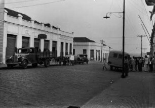 Straßenszene in Pirajuí : Eine Straßenszene in Pirajuí. Rechts und links der Straße Gebäude und Bürgersteige, sowie rechts Laternenmasten und Leitungen. Auf der Kopfsteinpflasterstraße parken Fahrzeuge, rechts auf dem Bürgersteig eine Personengruppe.