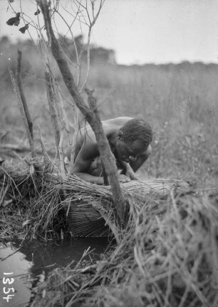 Am Wasser : Ein Mann an einem geflochtenen Gegenstandt. Möglicherweise stellt er seine Arbeit soeben fertig