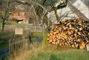 Ortseingang; Straße, Ortseingangsschild, Staketenzaun, Holzstapel