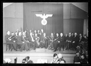 Musikkapelle auf Bühne, dahinter Fahne mit Reichsadler, im Vordergrund Publikum
