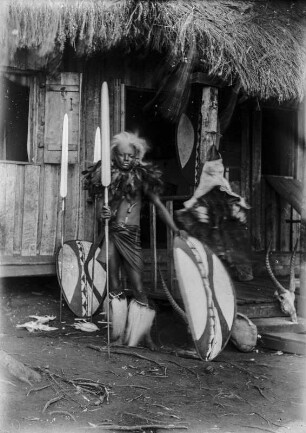 Krieger der Massai : Portrait eines Kriegers der Massai mit Speer und Schild vor einem Haus stehend