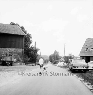 Tralau: ausgebauter Feldweg: in der Mitte Kinder mit Kinderkarre: vorn rechts Pkw: dahinter Neubau Einfamilienhaus: daneben Baumaterial: links Bauernhaus und Bäume: im Hintergrund Pkws und Haus