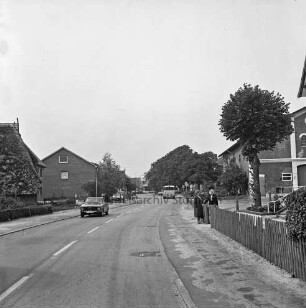 Alte Dorfstraße: vorn rechts Zaun: daran zwei Frauen: hinten Bauernhäuser und Grünanlage
