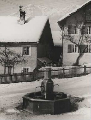 Oberbayern im Winter. Motiv aus Partenkirchen. Verschneite Häuser und ein wasserspeiender Brunnen an der Straße