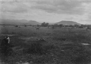 Steppe : Steppenlandschaft im Kilimandscharogebiet. Im Hintergrund erheben sich mehrere Hügel und in der Bildmitte liegt ein Koffer mit Utensilien. Am linken Bildrand sitzt ein Mann