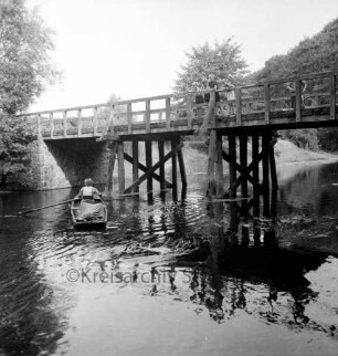 Trave: darüber Friedensbrücke: rechts oben Kinder am Geländer: daneben Mann mit Fahrrad: links Frau in Ruderboot: hinten Bäume