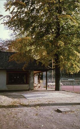 Tennis- und Hockey-Club Blau-Weiß e.V.: Kurpark: links Vereinsgebäude: davor Tische, Stühle und Baum: rechts hinten Tennisplatz