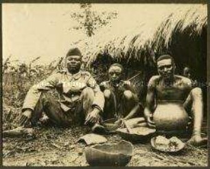 Askari und Afrikaner mit traditionellem Geschirr vor einer Hütte