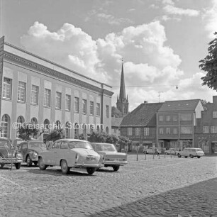 Marktplatz: vorn links und hinten rechts parkende Pkws: dahinter links Postgebäude, rechts Hindenburgstraße mit Wohn- und Geschäftshäusern: im Hintergrund Mitte Turm der Peter-Paul-Kirche