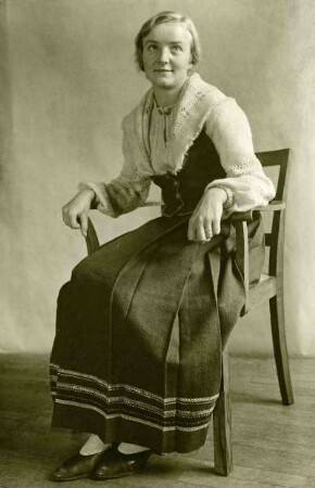 Frau sitzend auf hölzernen Stuhl, in Volkstracht mit handgewebtem Kleid.. Halbtotale