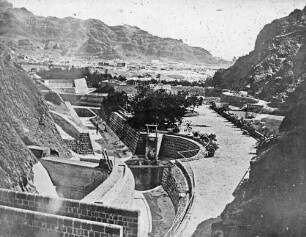 [Aden. Wasserleitung. Blick auf eine eng bebaute Siedlung an einer Bucht, vorne eingefasster Wasserlauf]