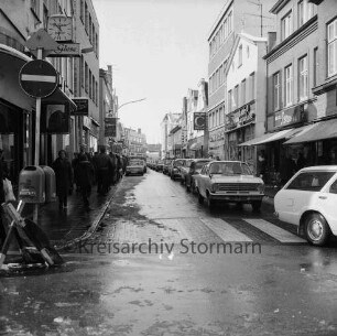 Hindenburgstraße / Mühlenstraße: vorn Zebrastreifen: rechts wartende Pkws: links Fußgänger: rechts und links Wohn- und Geschäftshäuser mit Feinkostgeschäft Peters, Landesbausparkasse, Commerzbank, Pelzgeschäft Günter Ludwig, Gardinen- und Bettengeschäft Meincke Hansen: 29. März 1975