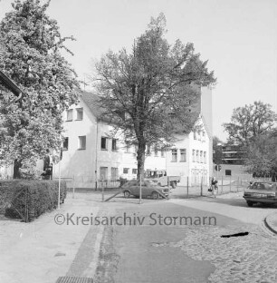 Feuerwache Woldenhorn: Schäferweg: [ehemaliges Elektrizitätswerk]: Bildmitte Feuerwehrgerätehaus, davor parkende Autos, Zaun: rechts dahinter Schlauchturm: auf Straße Fußgängerin, Auto: links Vorgarten, Baum, Hecke
