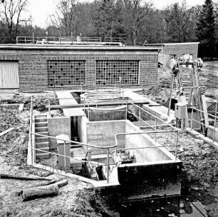Abwasser: Klärwerk: Wöhrendamm: 3. Bauabschnitt: Schmutzwassereinläufe: Becken im Rohbau mit Geländer und ersten Einbauten: rechts Förderband: hinten Maschinen- und Betriebsgebäude im Rohbau: im Hintergrund Bäume