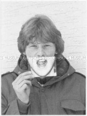 Männlicher Jugendlicher hält sich vor seinen Mund ein Foto von einem aufgerissenen Mund (Altersgruppe 14-17)