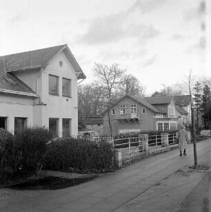 Hauptstraße: hinten rechts Frau auf Gehweg: daneben kleiner Baum: hinten Wohnhäuser mit Vorgärten: dazwischen VW-Bus und Bäume