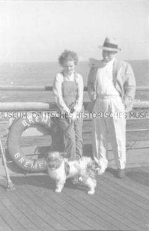 Junge Frau mit Hund neben rauchendem Herrn an Deck des Hochseepassagierdampfers "Bremen", neben einem Rettungsring an der Reling lehnend