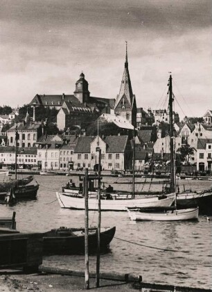 Blick auf Flensburg am Ostufer der Förde. Im Vordergrund ankernde Fischerboote. In der Bildmitte der Turm der um 1284 im gotischen Stil errichteten Marienkirche. Daneben das Gebäude des alten Gymnasiums, gegründet 1566