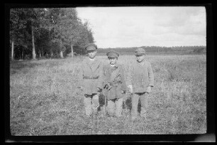 3 Jungen mit Schirmmützen und kurzen Mänteln auf einer Wiese am Waldrand