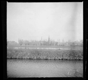 Comines-Warneton (Belgien): Ortsansicht mit Schornsteinen, Glockenturm und zwei Kirchtürmen, im Vordergrund ein Kanal und ein leicht überflutetes Feld ("Belgisch-Comines mit Kanal")