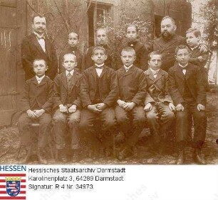 Knodt, Karl Ernst (1856-1917) / Porträt, mit Gruppe von Kindern vor Haus stehend, dabei bezeichnet 'Der Herr Vikar' und Sohn Theo[dor] (1891-1971), sitzend 1. Reihe 1. v. r.