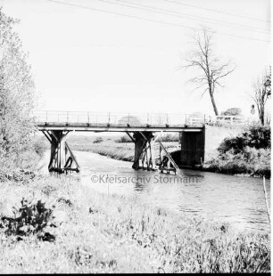 Trave: Holzbrücke in baufälligem Zustand: vorn Böschung: hinten rechts und links Bäume: dahinter Wiesen