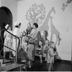 Erzieherinnen in einer Kinderkrippe (oder einem Kindergarten) mit Kindern und einer Mutter, die ihren kleinen Sohn auf dem Arm trägt
