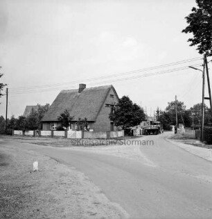 Straßenansicht: vorne eine Abzweigung, dahinter ein Wohngebäude, rechts ein Traktor mit Anhänger