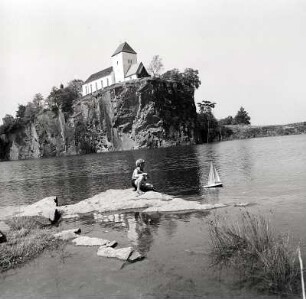See und Kirche : Beucha. Landschaftsbild mit einer Kirche auf einem Felsvorsprung an einem See. Im Vordergrund sitzt ein kleiner Junge auf einem Stein im See und schaut seinem kleinen Spielzeug Segelboot, das im Wasser schwimmt nach