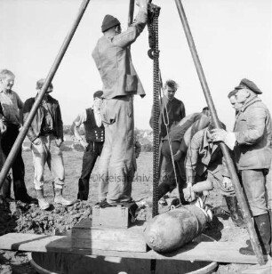 Bombenräumung: Wiese am Stadtrand: vorn Trichter mit Dreibein und Brett: darauf entschärfte 250-Kilogramm-Bombe: vorn Mitte Mann an Gestänge: dahinter mehrere Arbeiter des Bombenräumkommandos Kiel [später: Kampfmittelräumdienst Kiel]: hinten Wohnblocks mit Gärten