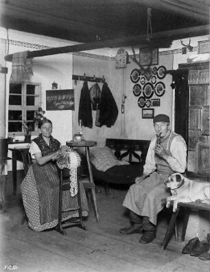 Gustav Hiemann mit Ehefrau in der Bauernstube : Steinbach im Erzgebirge, Bauernstube von Gustav Hiemann. Frau klöppelt, Bauer mit Hund auf der Ofenbank