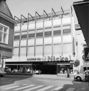 Nickel: Mühlenstraße / Lübecker Straße: Kaufhaus: Haupthaus Mühlenstraße, mit Firmenname und Leuchtschrift "Tschibo": im 2. Obergeschoss Restaurant: Fußgänger, Auto: im Vordergrund Einmündung der Hindenburgstraße, Fußgängerüberweg mit Zebrastreifen: Eckhaus links Delekatessengeschäft Peters