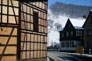 gekrümmte Straße mit Häusern, dahinter geht das Gelände steil bergan, oben Wald