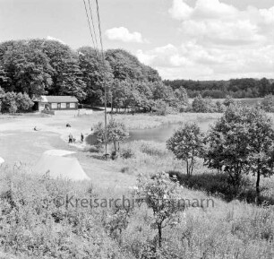 Lütjensee: Campingplatz am Nordufer: vorn Gebüsch und Bäume: hinten rechts See: links Zelt und Personen am Ufer: hinten links Holzhaus mit Gastronomie und Toiletten: dahinter Wald