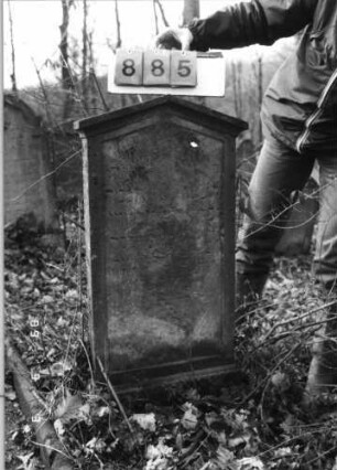 Grabstein 885 (Index: 99751189) Material: Sandstein Zustand: abgeblätt/verwitt/großer Textverlust Maße HxBxT: 89x48x8 cm Beschreibung: Stele, profiliert. Sockel, giebelart. Abschl. m. Verdach. , Schriftfeld eingetieft: Grabstein 885