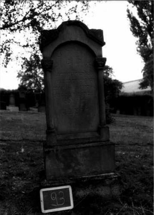 Grabstein 93 (Index: 100107259) Material: Sandstein Zustand: leicht verwittert, Aufsatz fehlt Maße HxBxT: 160x51x20 cm Beschreibung: ädikulaartige Stele mit Halbsäulen, getreppter Sockel, schräg eingezogener Rundbogenabschluß mit Verdachung, Schriftfeld mit Rundbogen Symbolik: Blattwerk, Rundbogenfries: Grabstein 93