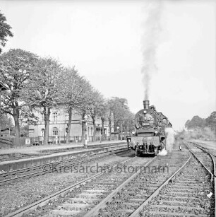 Bahnhofsgelände: Gleise mit Dampflokomotive: links Bahnsteige, Wasserkran, Baumreihe, Bahnhofsgebäude: Blick nach Norden